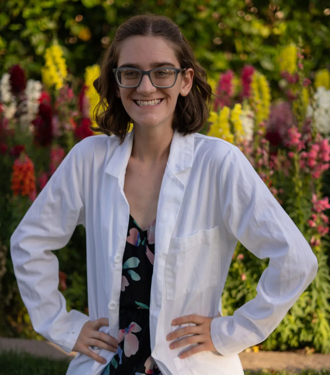 Young person wearing a lab coat smiles with her hands on her hips. Behind her are color spring flowers and greenery.