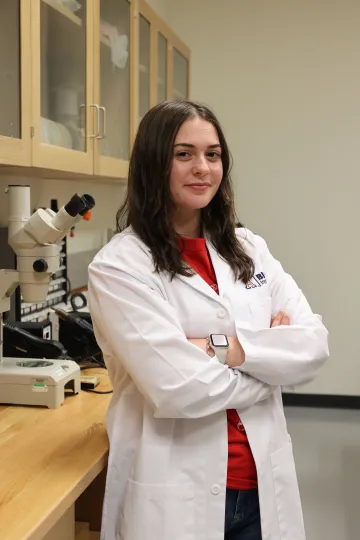Student in a white lab coat stands confidently with crossed arms in a laboratory setting, smiling at the camera, with a microscope and other scientific equipment in the background.